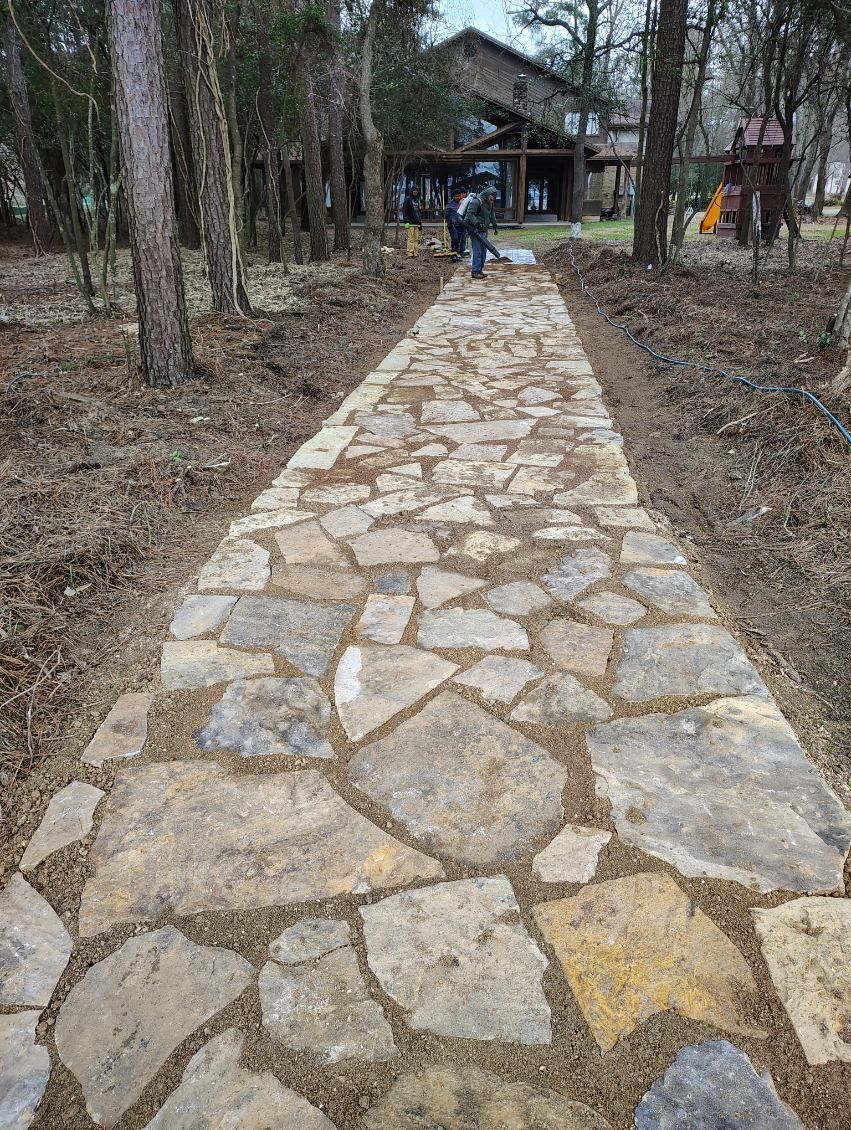 Stone pathway leads to a house through a wooded area. Man works on path in the distance.