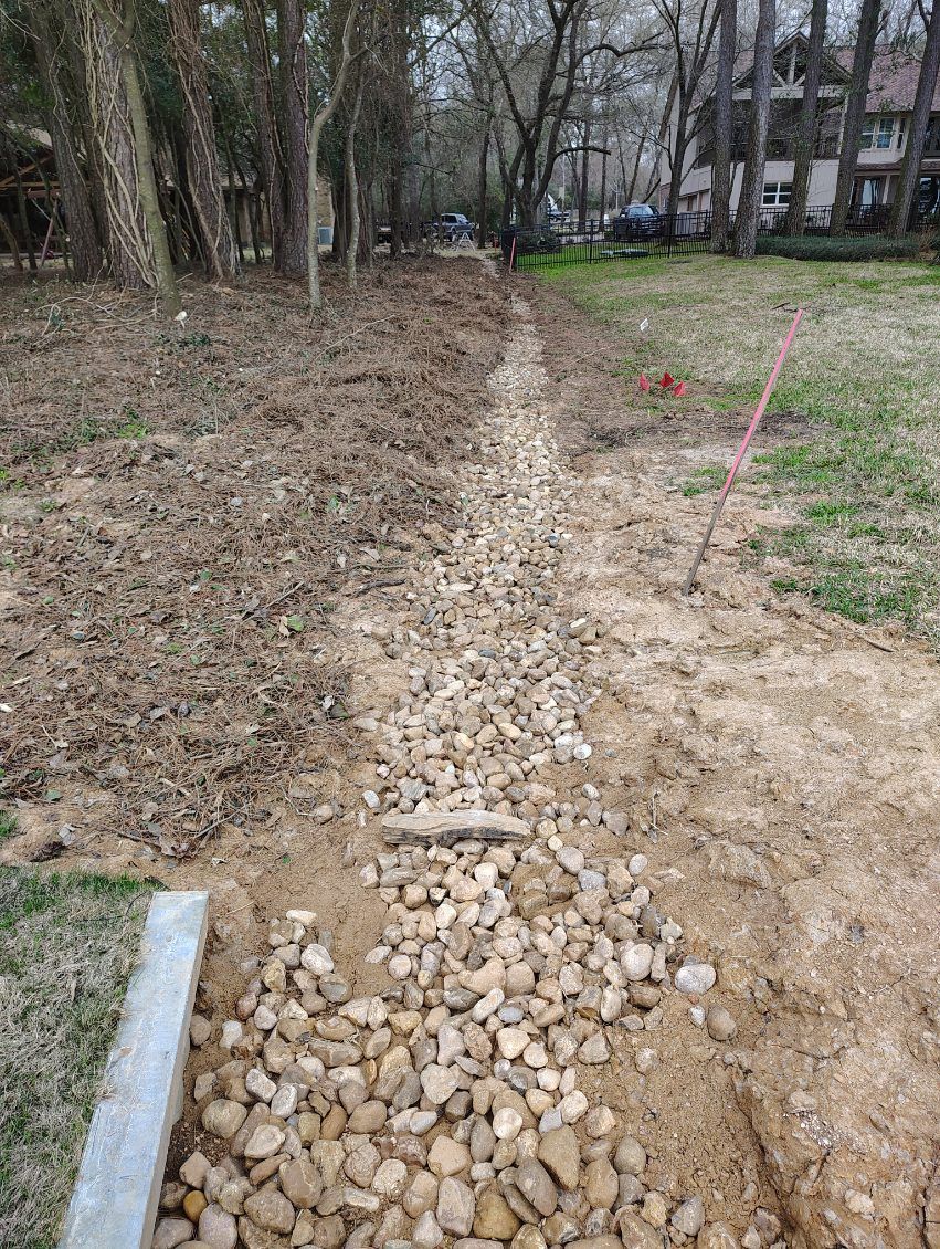 A gravel-lined trench in a yard, possibly for drainage, with dirt on either side and trees in the background.