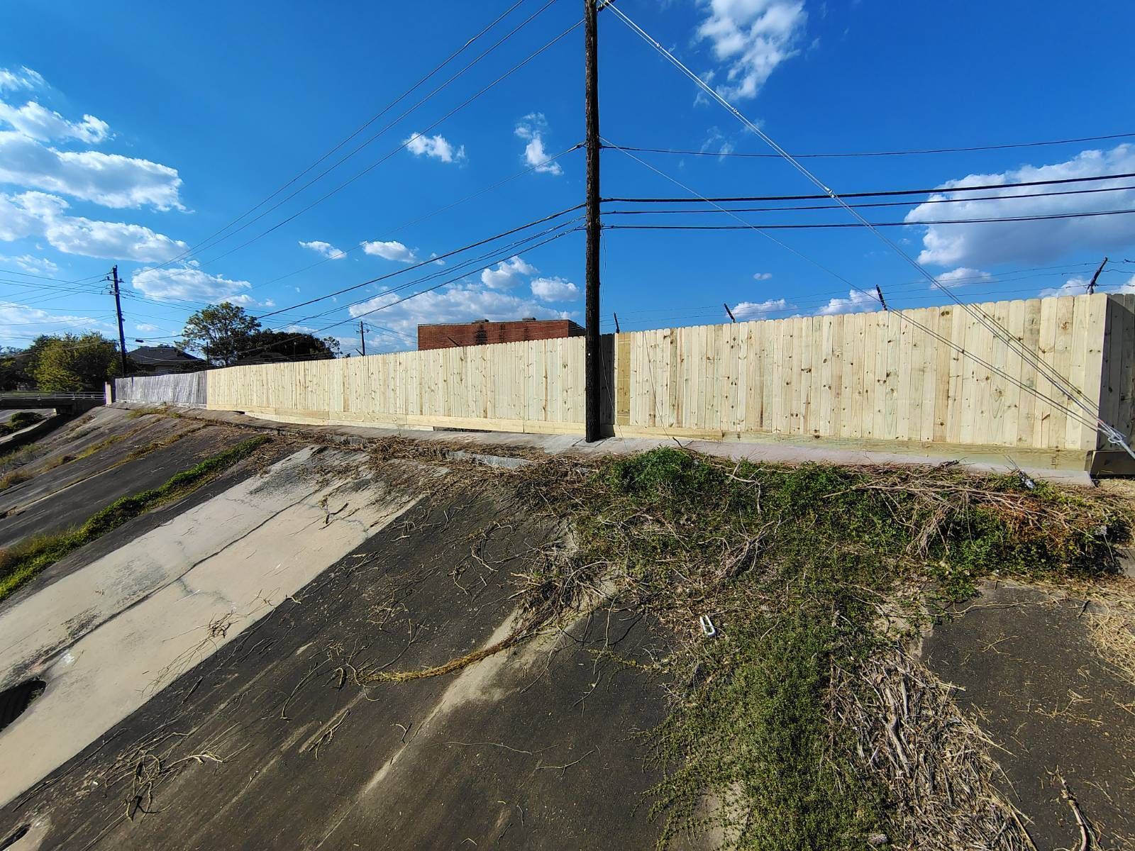 A long wooden fence sits atop a concrete embankment under a blue sky, with power lines overhead.