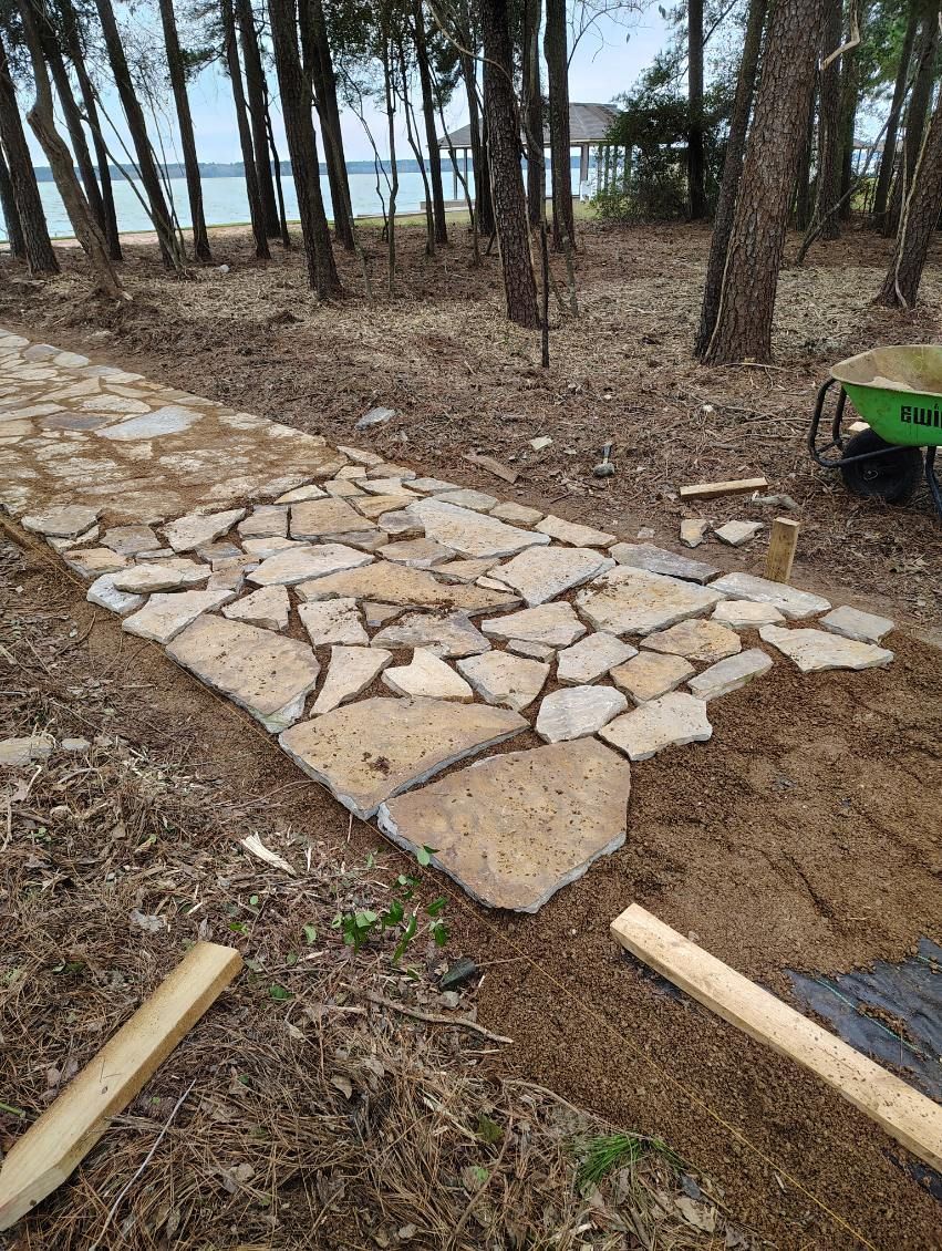 A pathway made of irregularly shaped stones, surrounded by soil and trees, leading to a body of water.