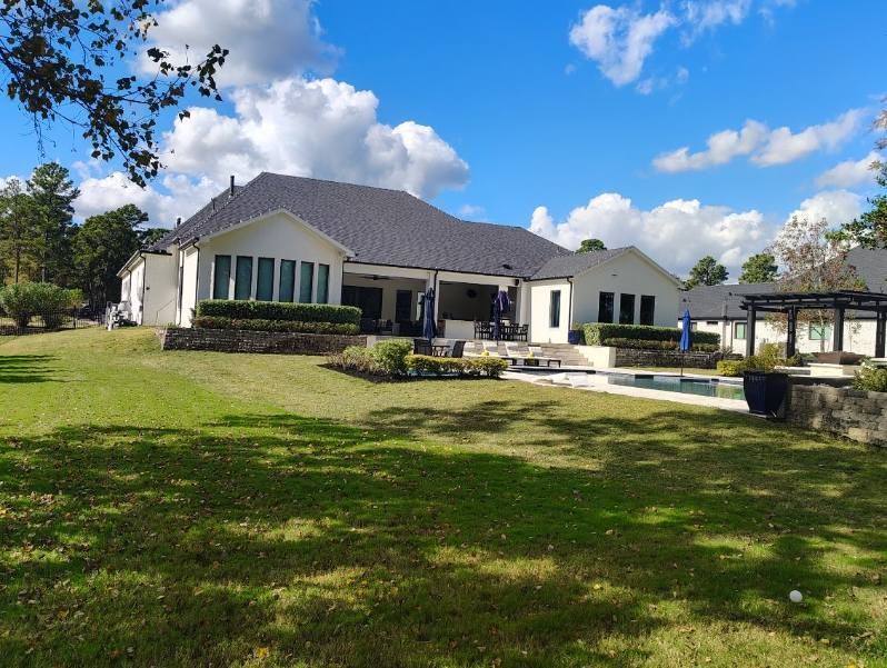 Backyard view of a white house with dark roof, pool, and green lawn under a partly cloudy blue sky.