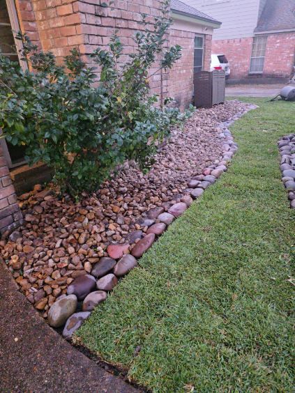 Landscape with a strip of green grass, a rock bed, and a shrub next to a brick building.