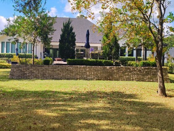 A large house with a stone wall in front, on a sunny day. Green grass and trees frame the scene.