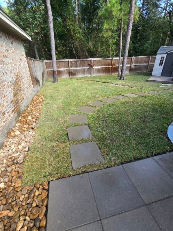 Backyard with stone path through grass, bordered by brick wall and wooden fence, shed in the distance.