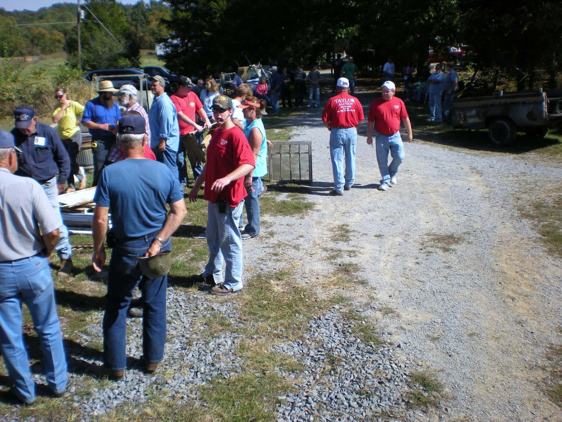 Group of people gathered on a gravel road, some in red shirts. Outdoors, sunny day.