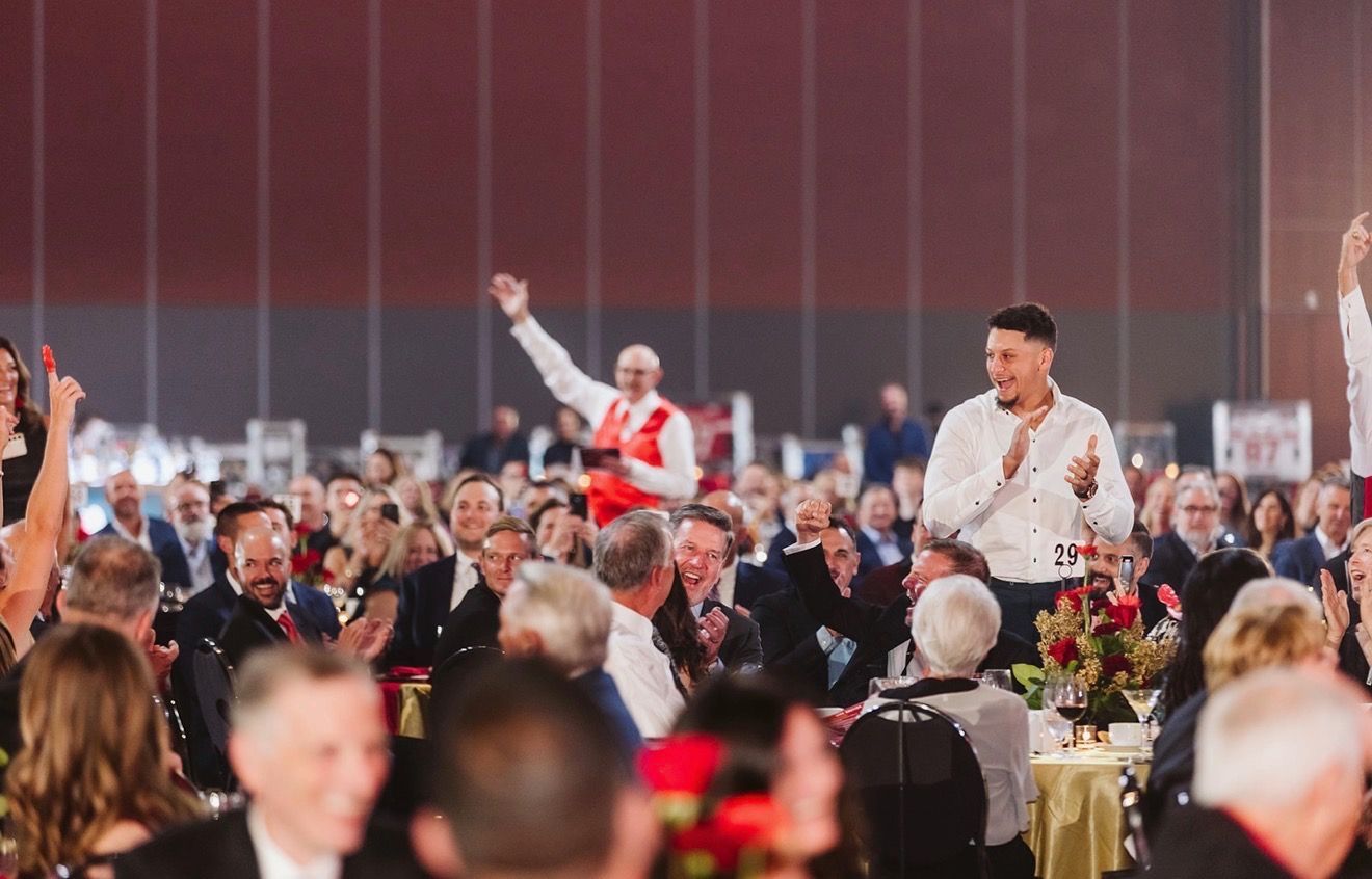Audience applauds as two men stand at a gala. One waves. Red and gold decor, indoor setting.