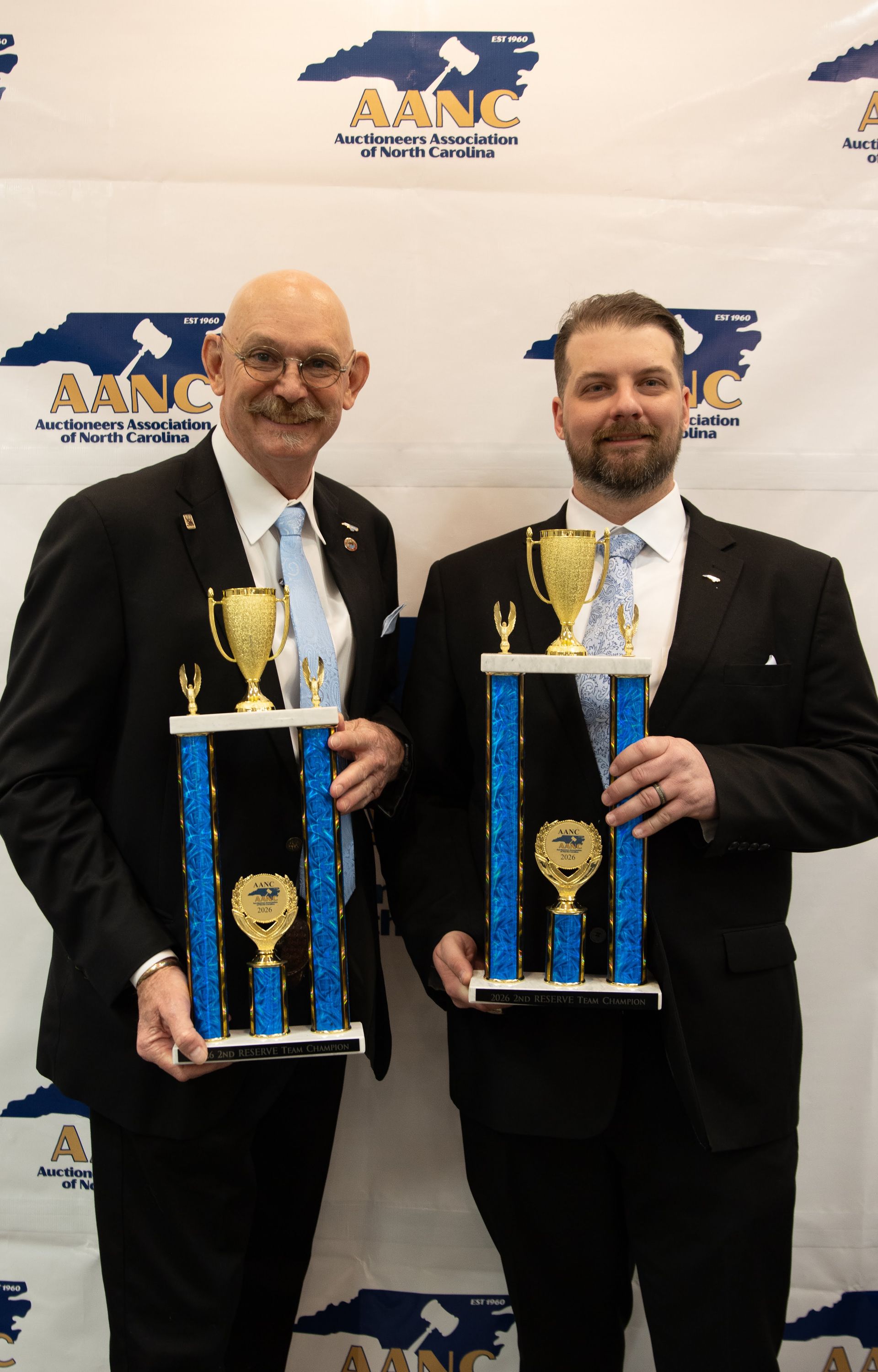 Two men in suits holding trophies in front of a banner.