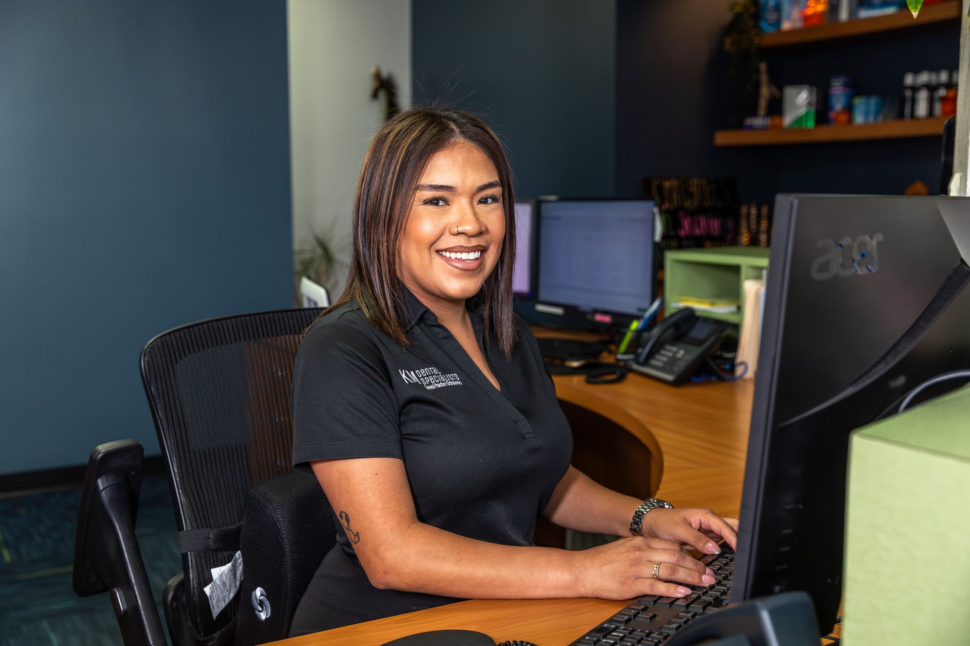 Woman smiling at desk, typing on keyboard. Blue wall, computer monitors, and office setting.