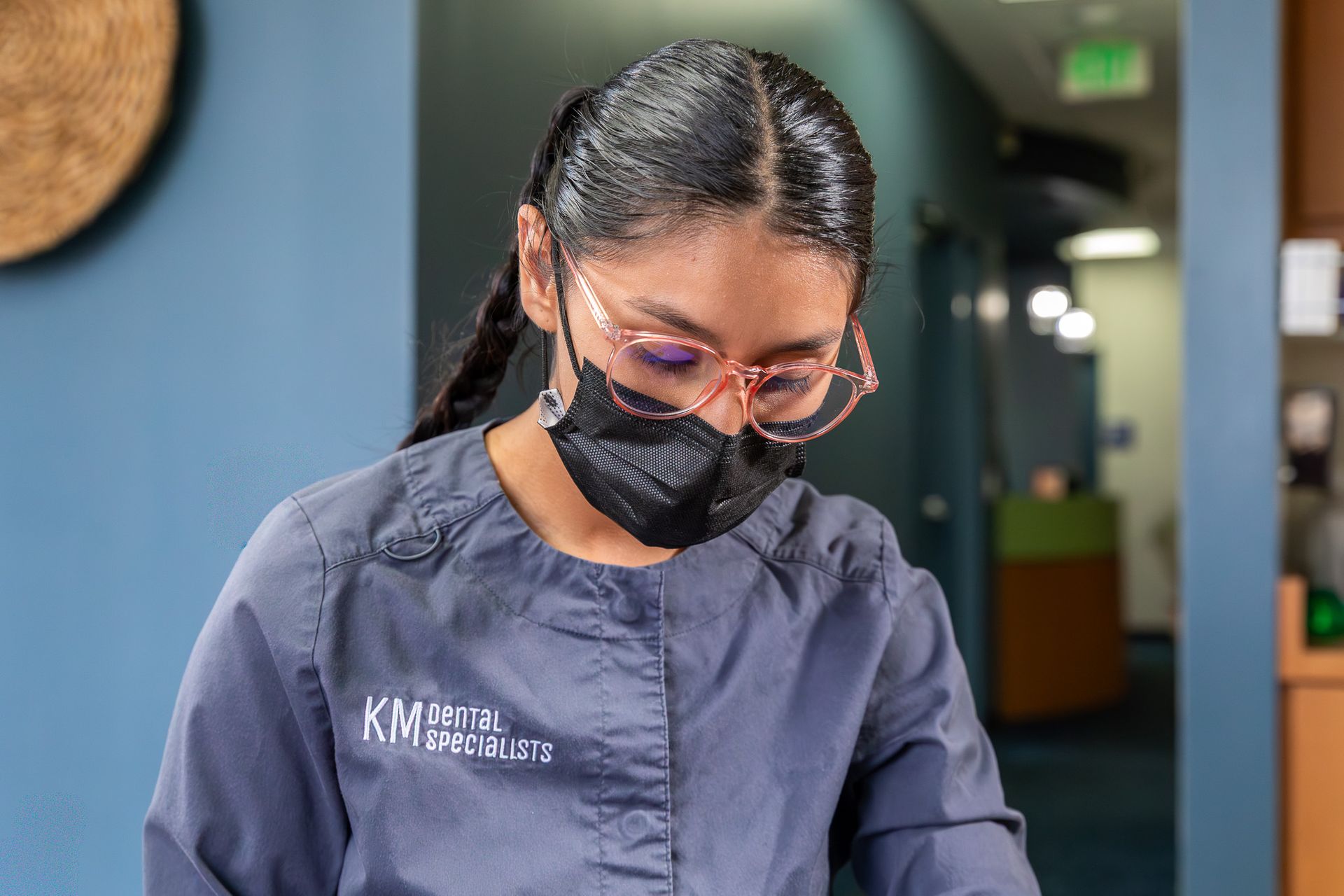 Dental assistant in a blue uniform and black mask, looking down.