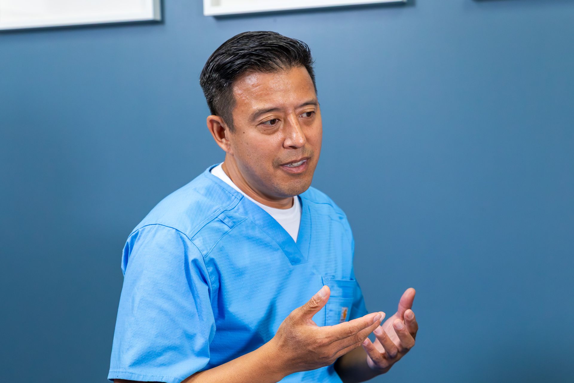Man in blue scrubs gesturing, speaking; blue wall background.