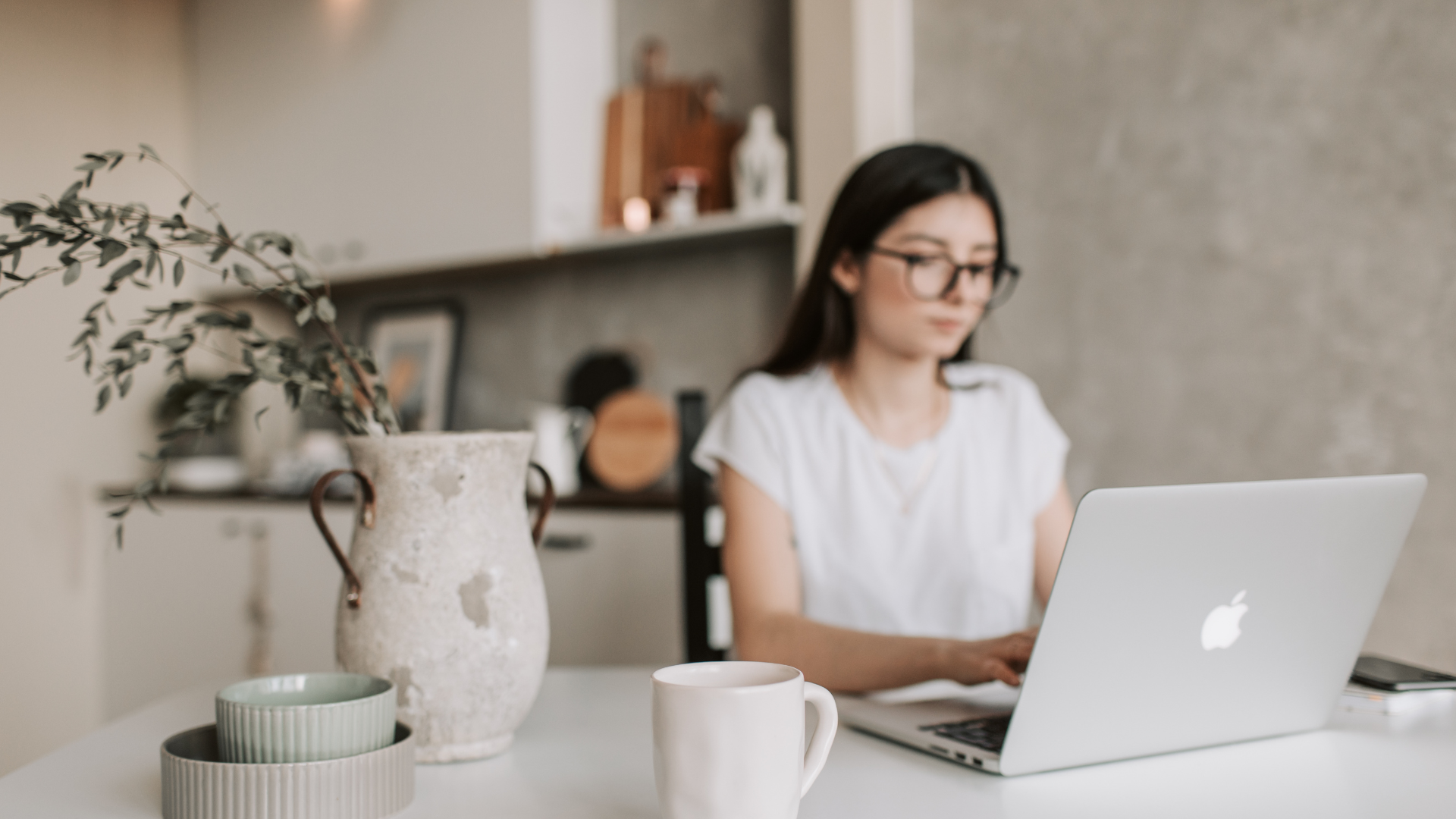 Woman with glasses working on a laptop at a table in a home setting, with a coffee mug and vase.