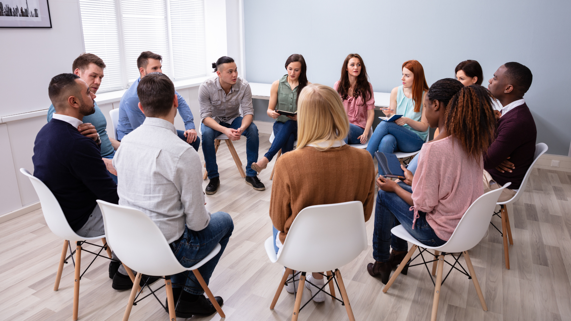 Group therapy session: Diverse people sit in a circle, talking and listening. Bright room.