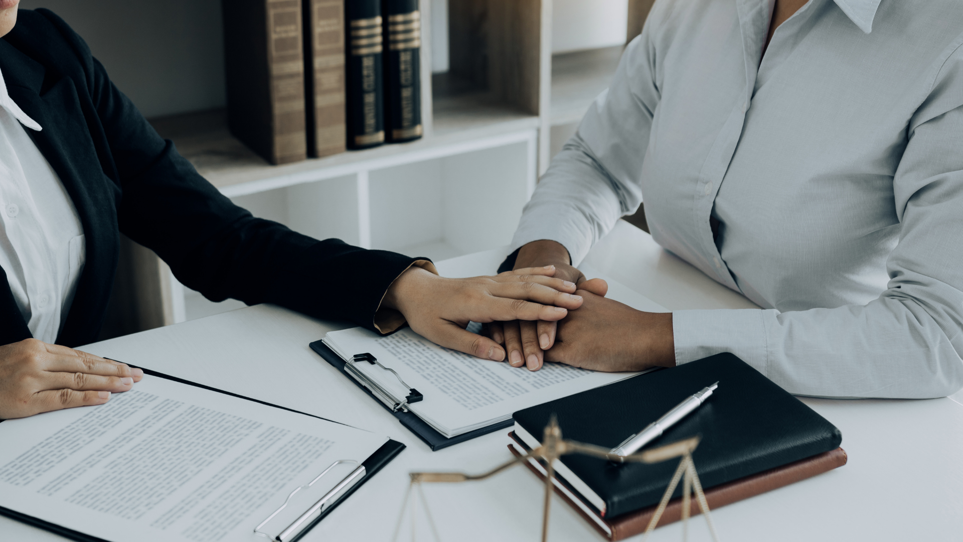 Lawyer comforting a client, hands clasped, at a desk with documents and law books in a professional setting.