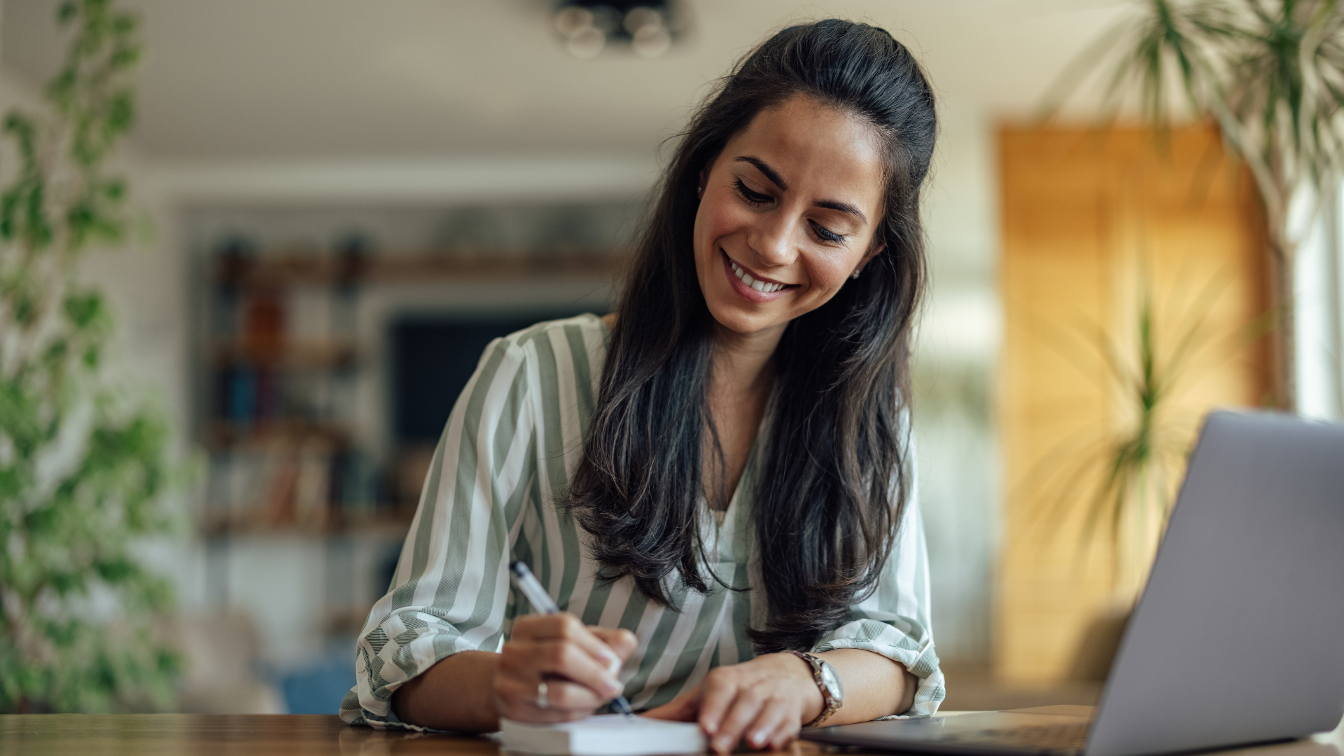 Woman with dark hair smiles while writing at a table with a laptop, bright room.