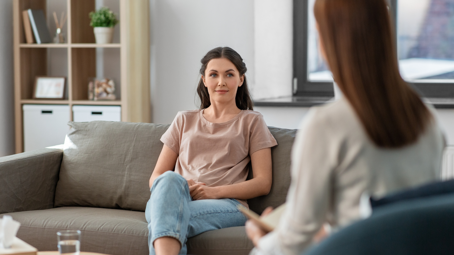 Woman in casual clothes seated on a couch, facing a person seated and holding a notepad in a counseling session.
