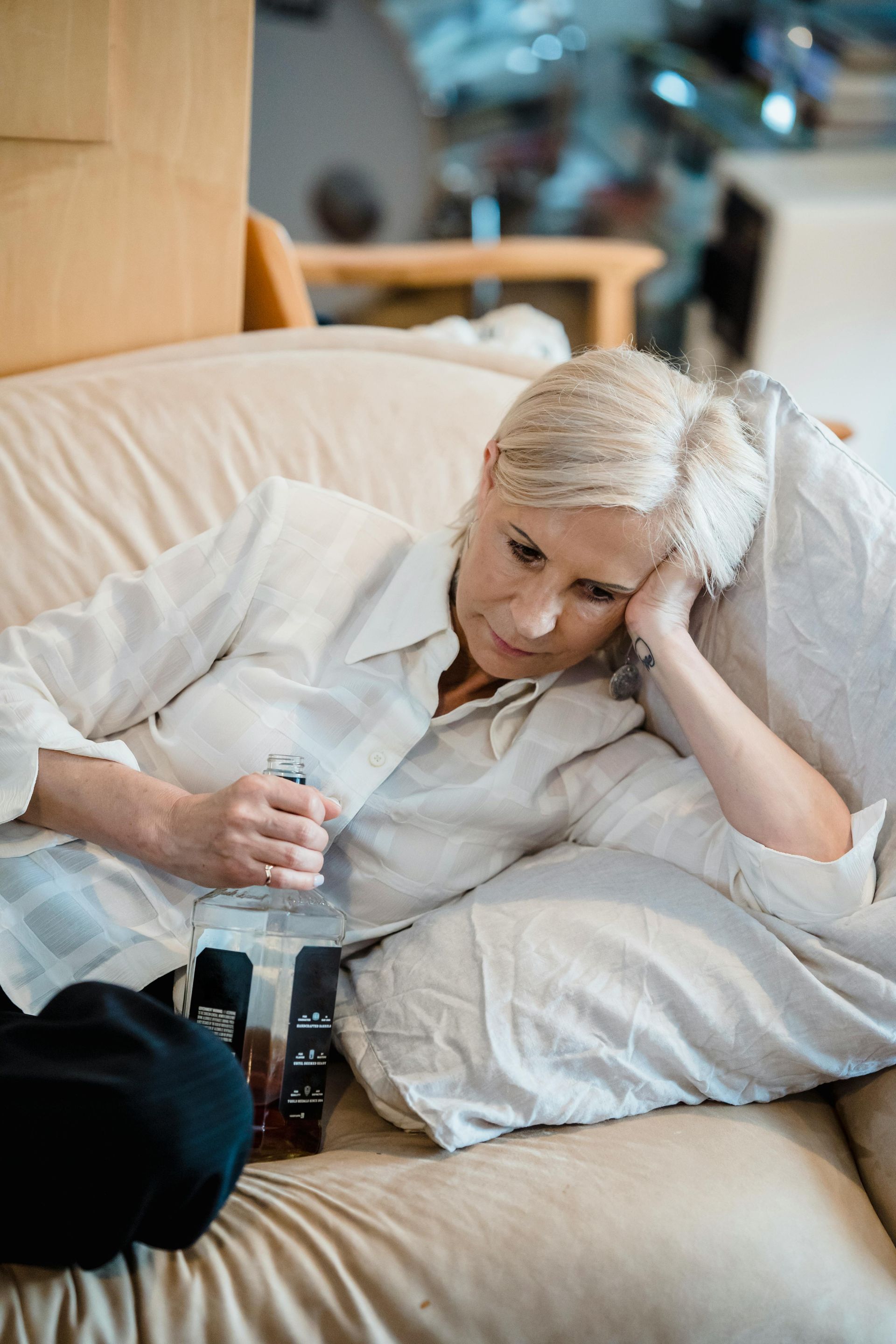 Woman lying on a couch, holding a liquor bottle, looking down with a sad expression.