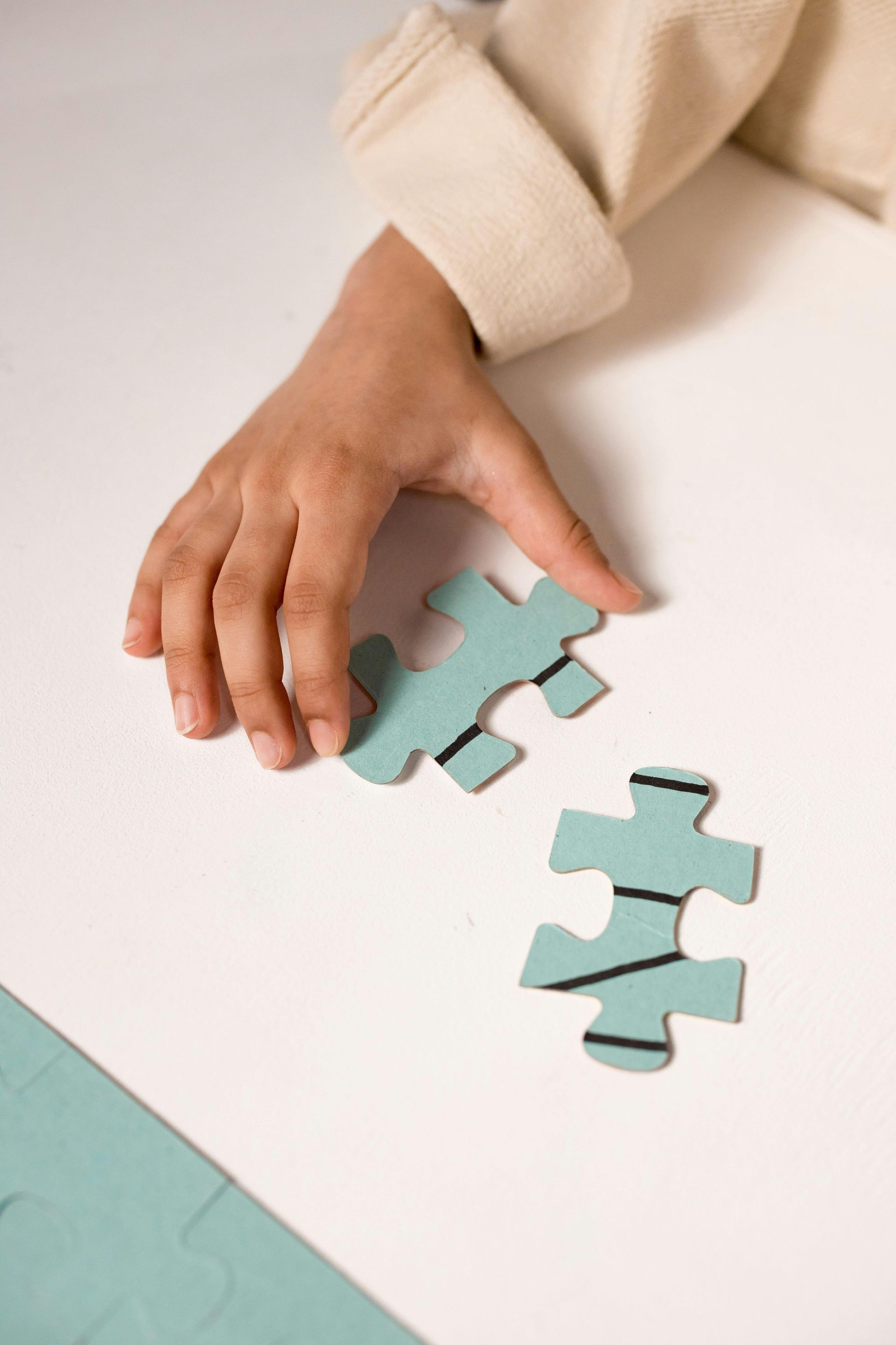 Child's hand holding a blue puzzle piece, about to connect it with another piece on a white surface.