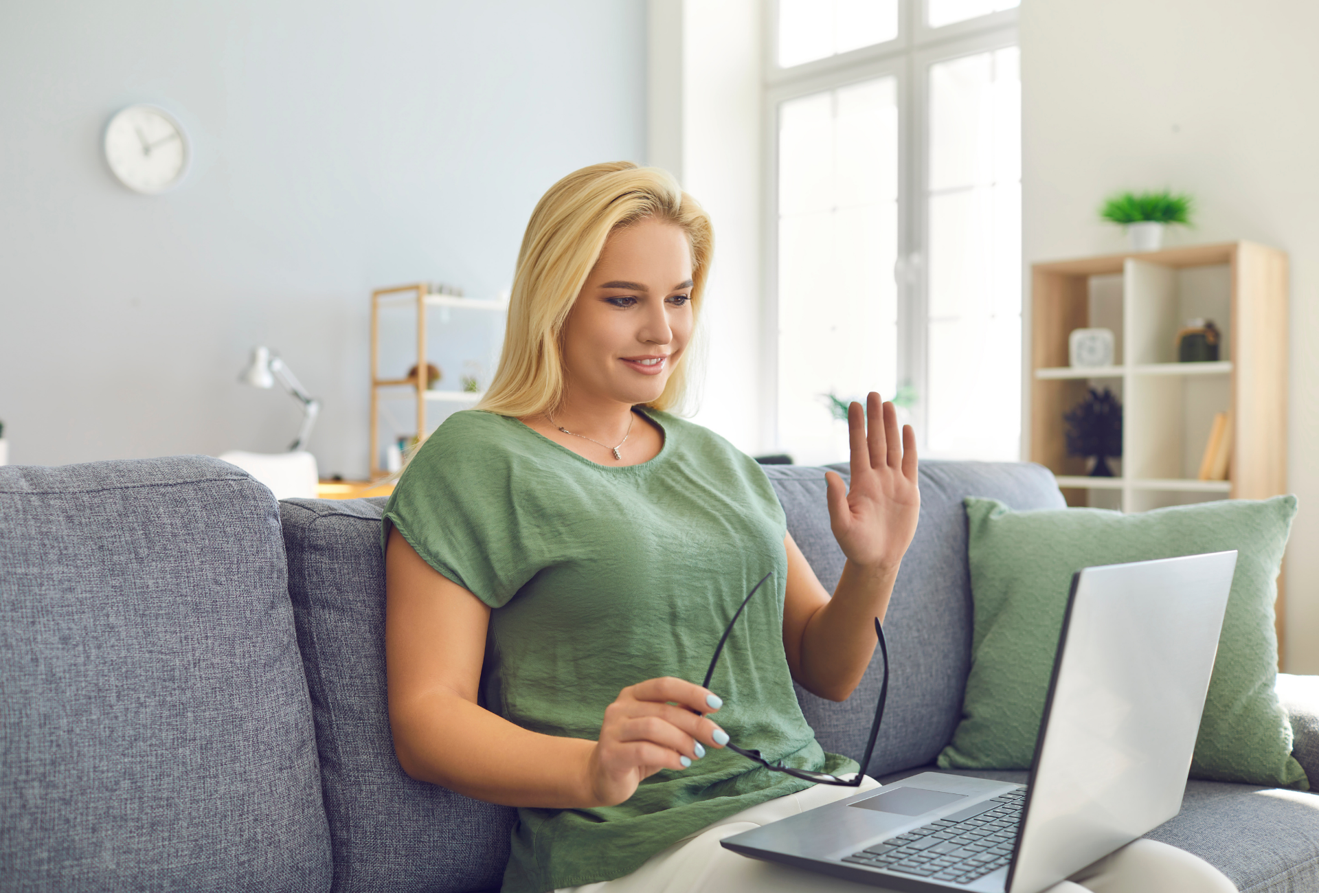 Blonde woman waves from a sofa during a video call on her laptop. Green shirt, bright room.