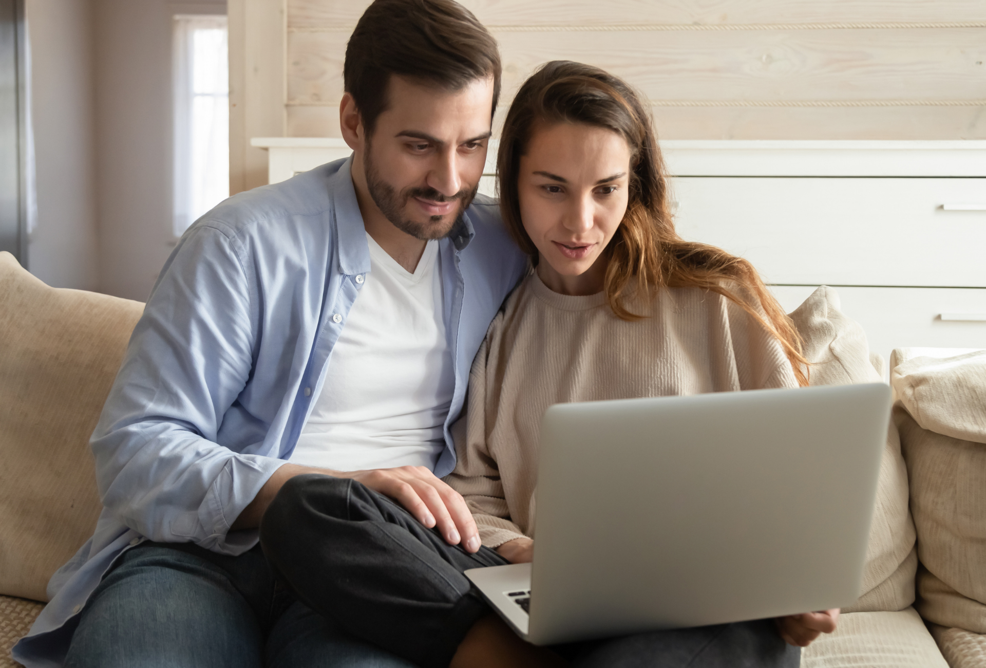 Couple on a couch looking at a laptop together, focused and surprised, in a living room.