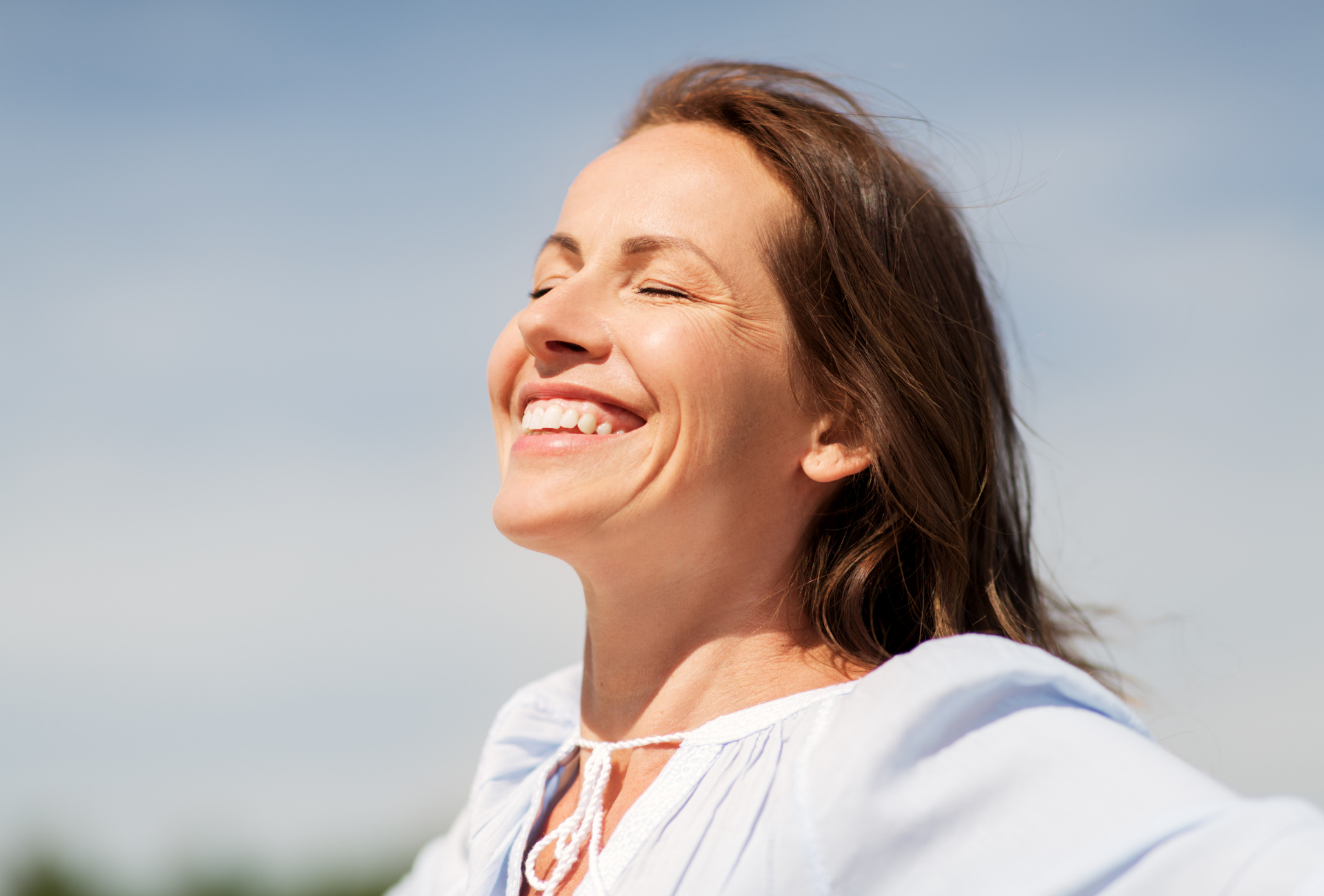 Woman with eyes closed and a wide smile, outdoors under a blue sky, enjoying the sun.