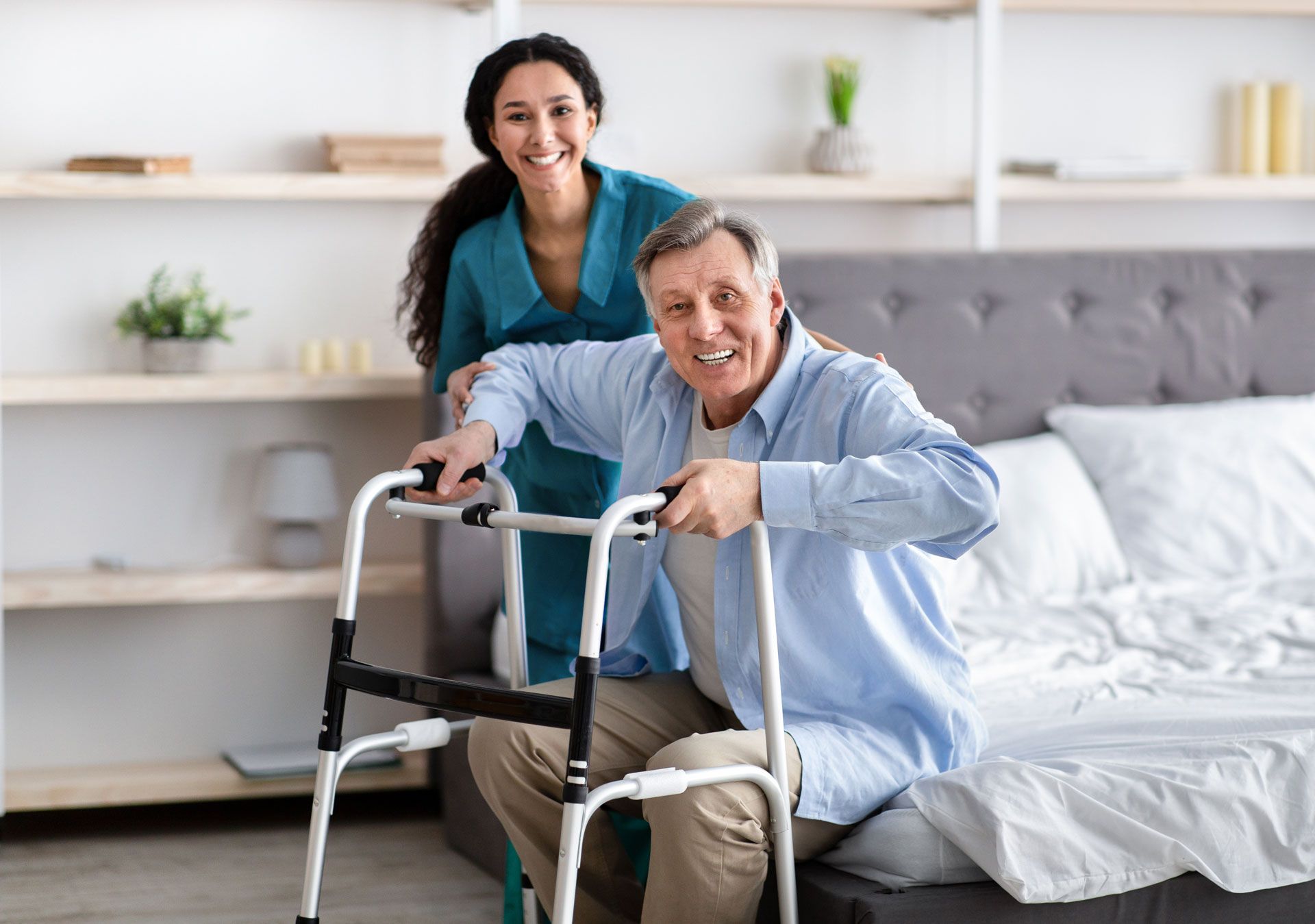 A smiling healthcare worker assists an elderly man using a walker in a bedroom. They are looking at the camera.