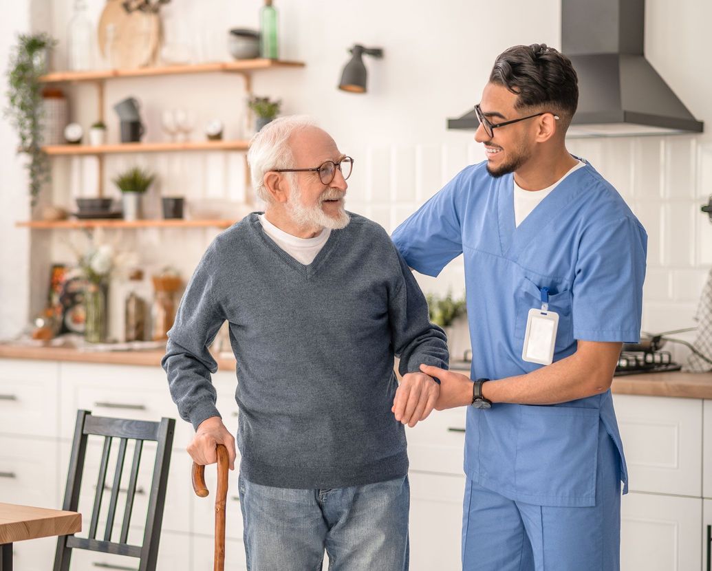 A young male caregiver in blue scrubs assists an elderly man with a cane in a home kitchen.