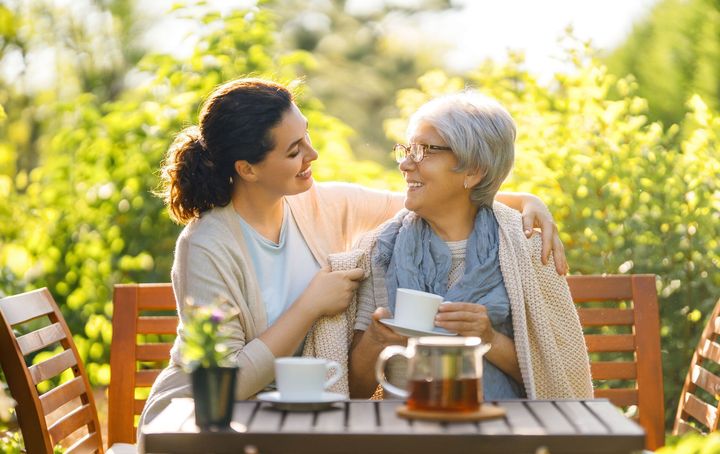 A young woman embraces an older woman while both smile, sitting at a table outdoors with tea.