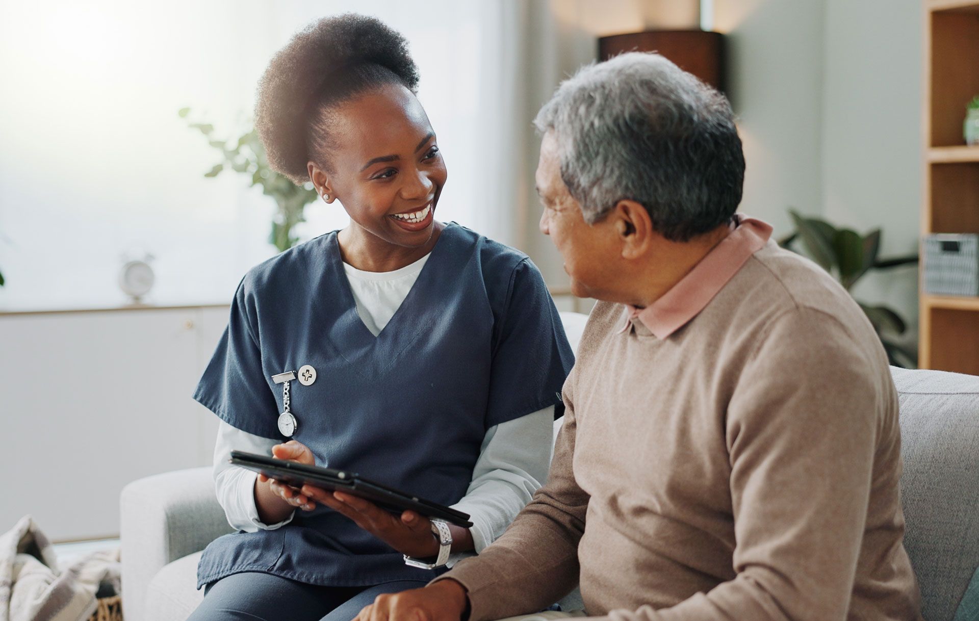 A Black female nurse in blue scrubs smiles while showing a tablet to an older man seated on a couch. They are indoors.