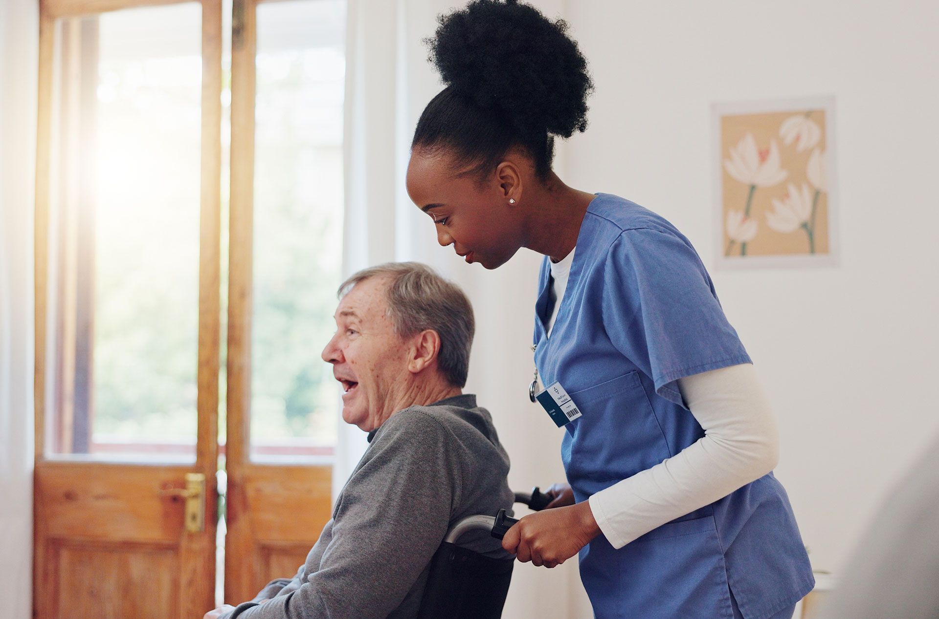 A Black nurse in blue scrubs pushes an elderly man in a wheelchair inside a home. He appears surprised or alarmed.