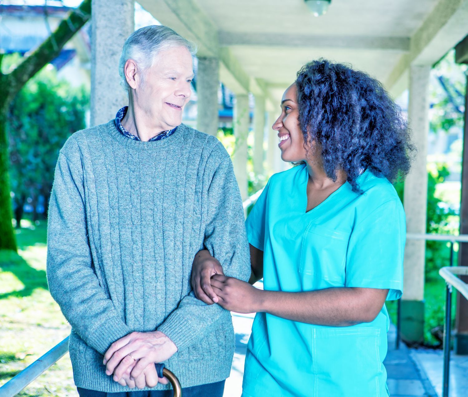 A young nurse smiles at an elderly woman in a wheelchair. They are indoors, and the woman is looking up at the nurse.