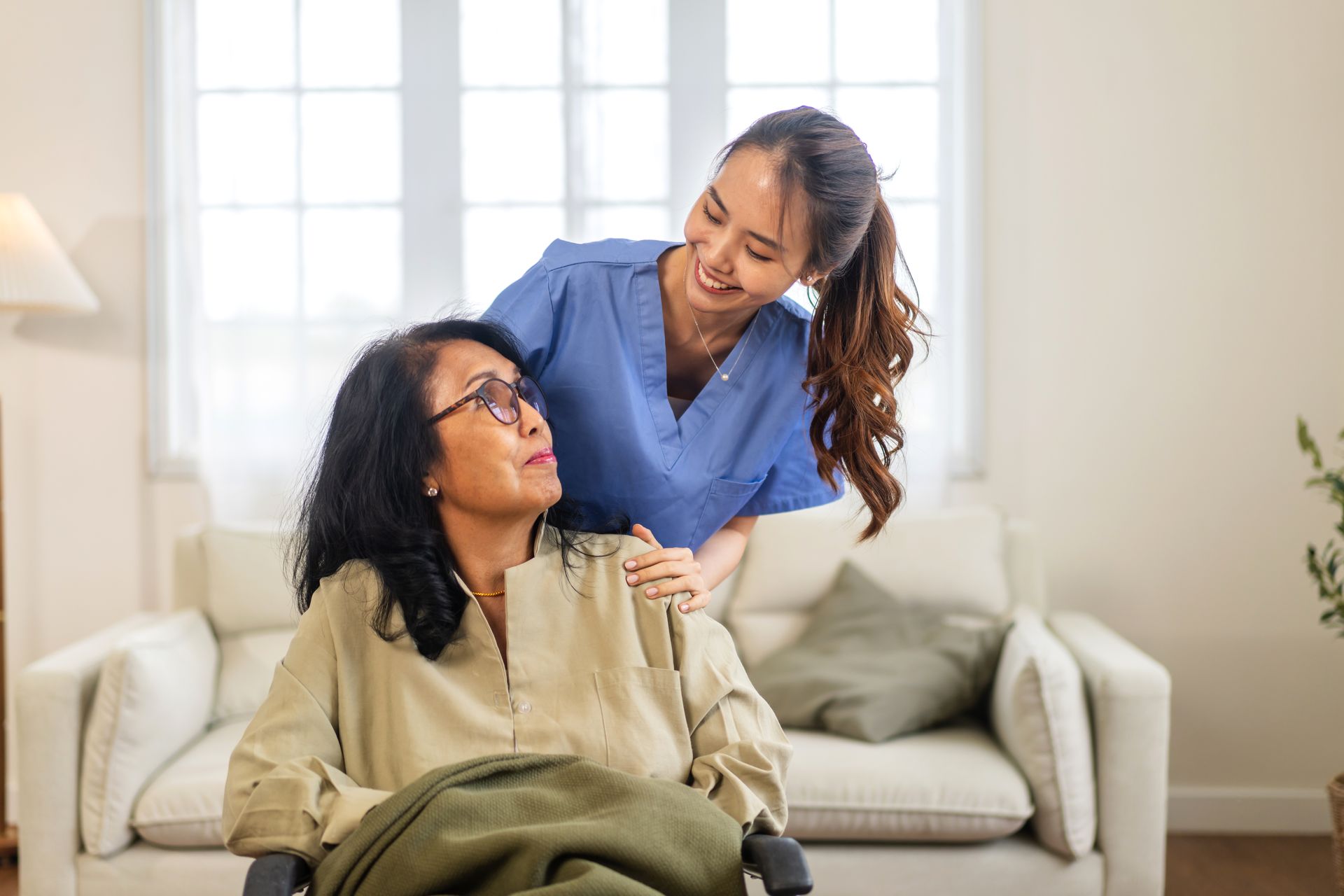 Caregiver leaning over seated woman in a bright living room, speaking gently and resting a hand on her shoulder