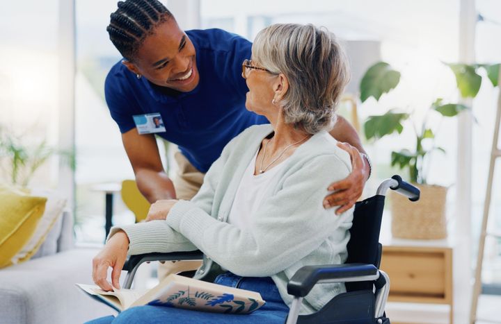 Caregiver in blue uniform smiling at an elderly woman in a wheelchair in a bright home setting