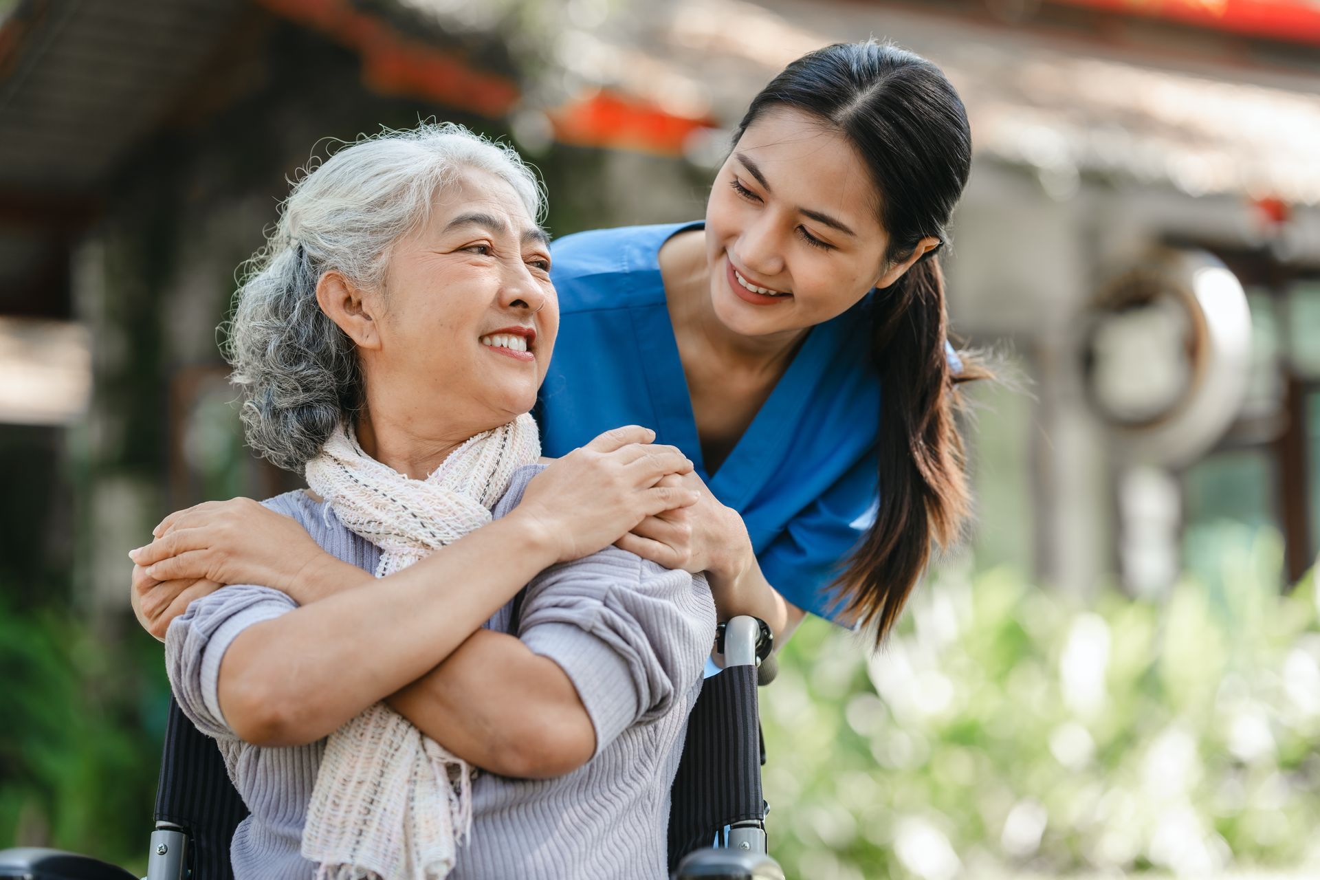 Two smiling women embracing outdoors, one seated and one leaning in with a hand on her shoulder