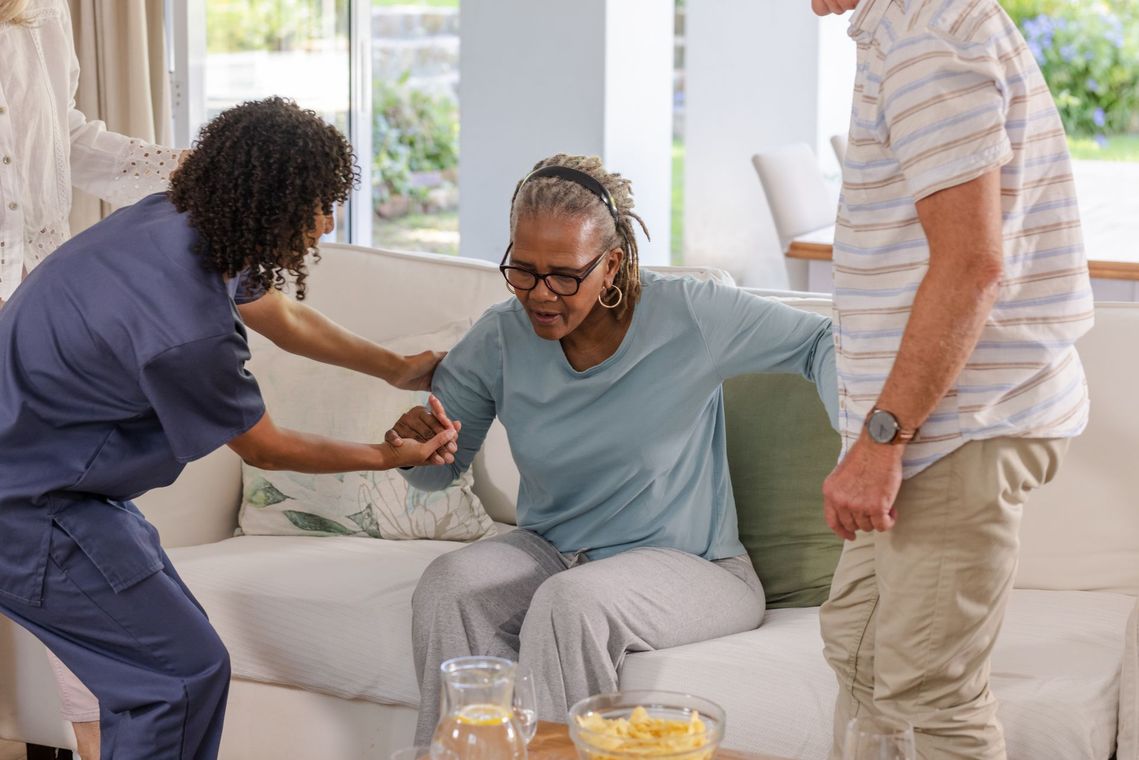 Caregiver helping a seated woman in a bright living room while another person stands nearby
