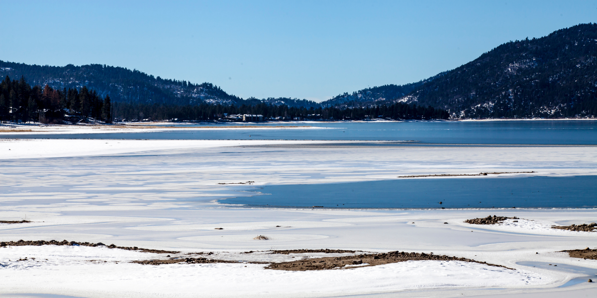 A partially frozen Big Bear Lake surrounded by snow-covered mountains under a clear blue sky.