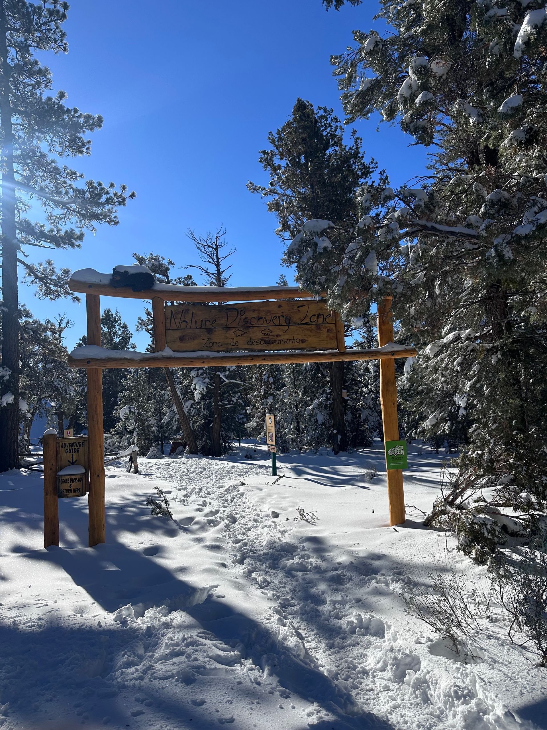 A wooden entrance arch covered in snow on a sunny day in a snowy, evergreen forest.