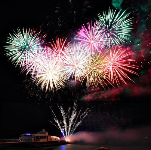Fireworks erupt over a pier, displaying colorful bursts against a dark sky.