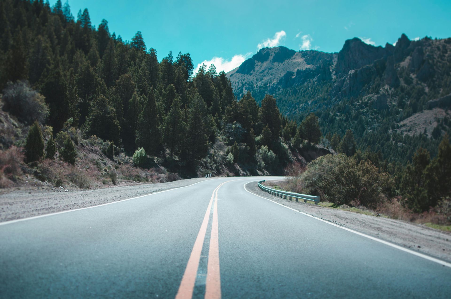 Paved road winding through a mountain landscape, lined with evergreen trees under a bright blue sky.