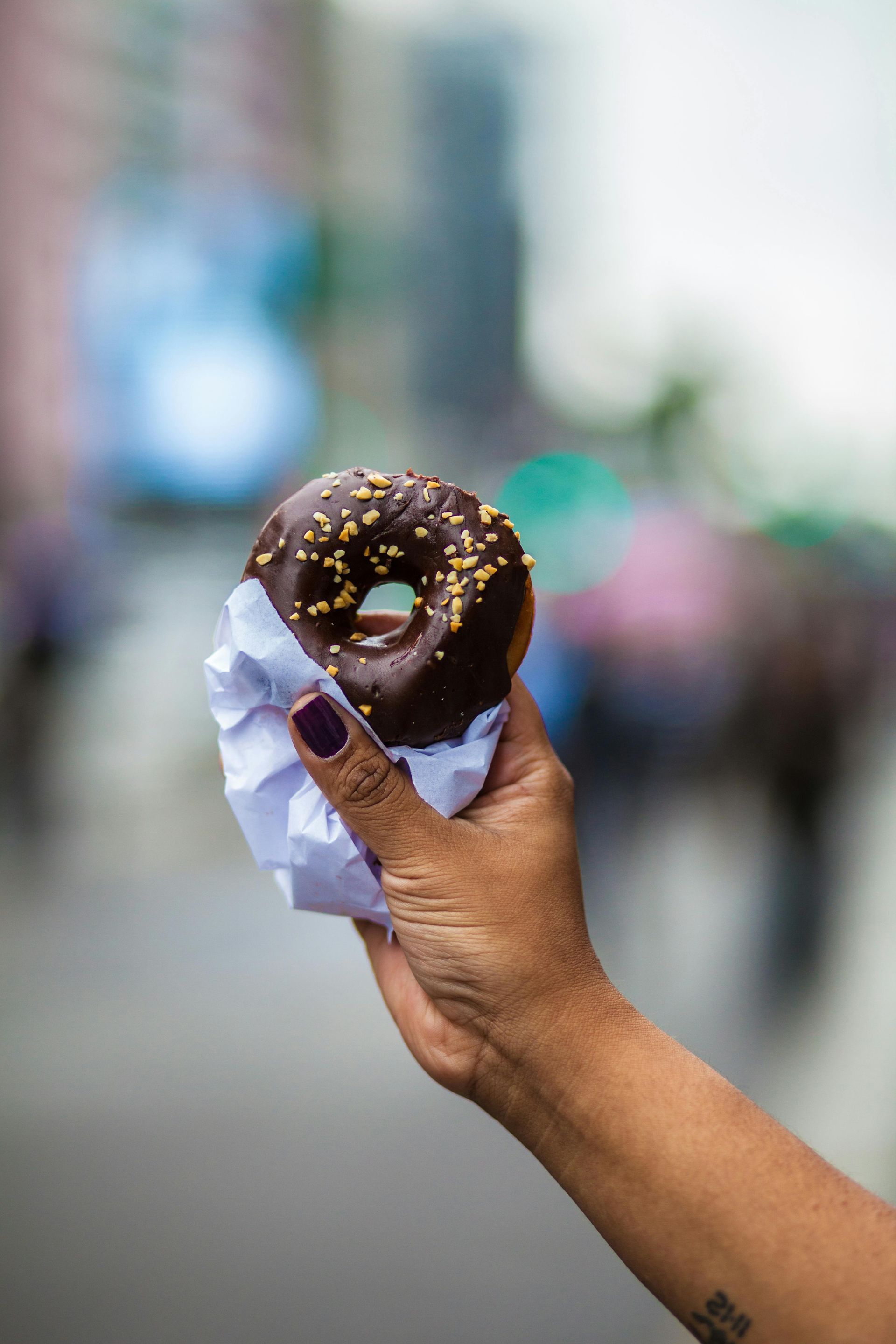 Hand holding a chocolate frosted donut with sprinkles, wrapped in white paper, blurred street background.