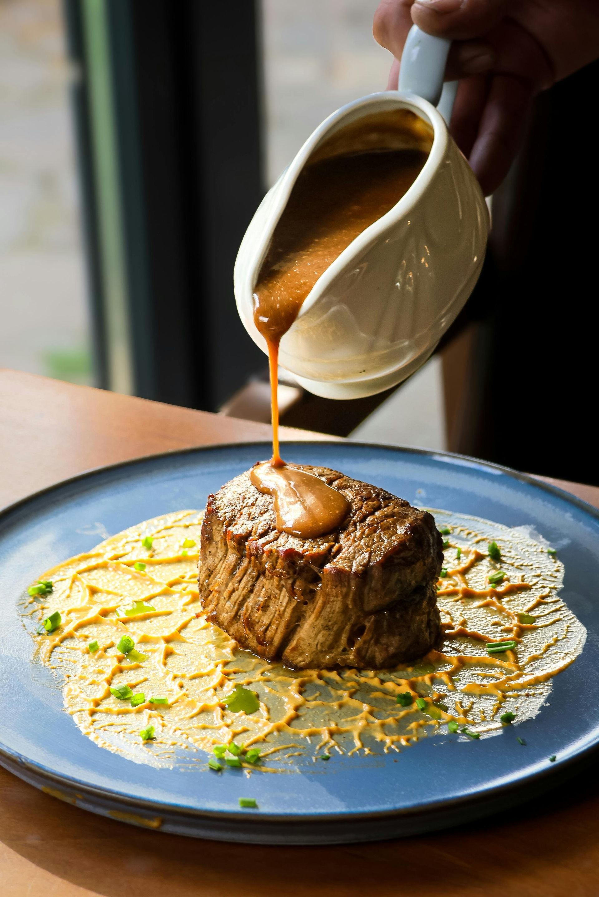 Filet mignon steak being drizzled with sauce on a blue plate, with a cream-colored sauce boat.