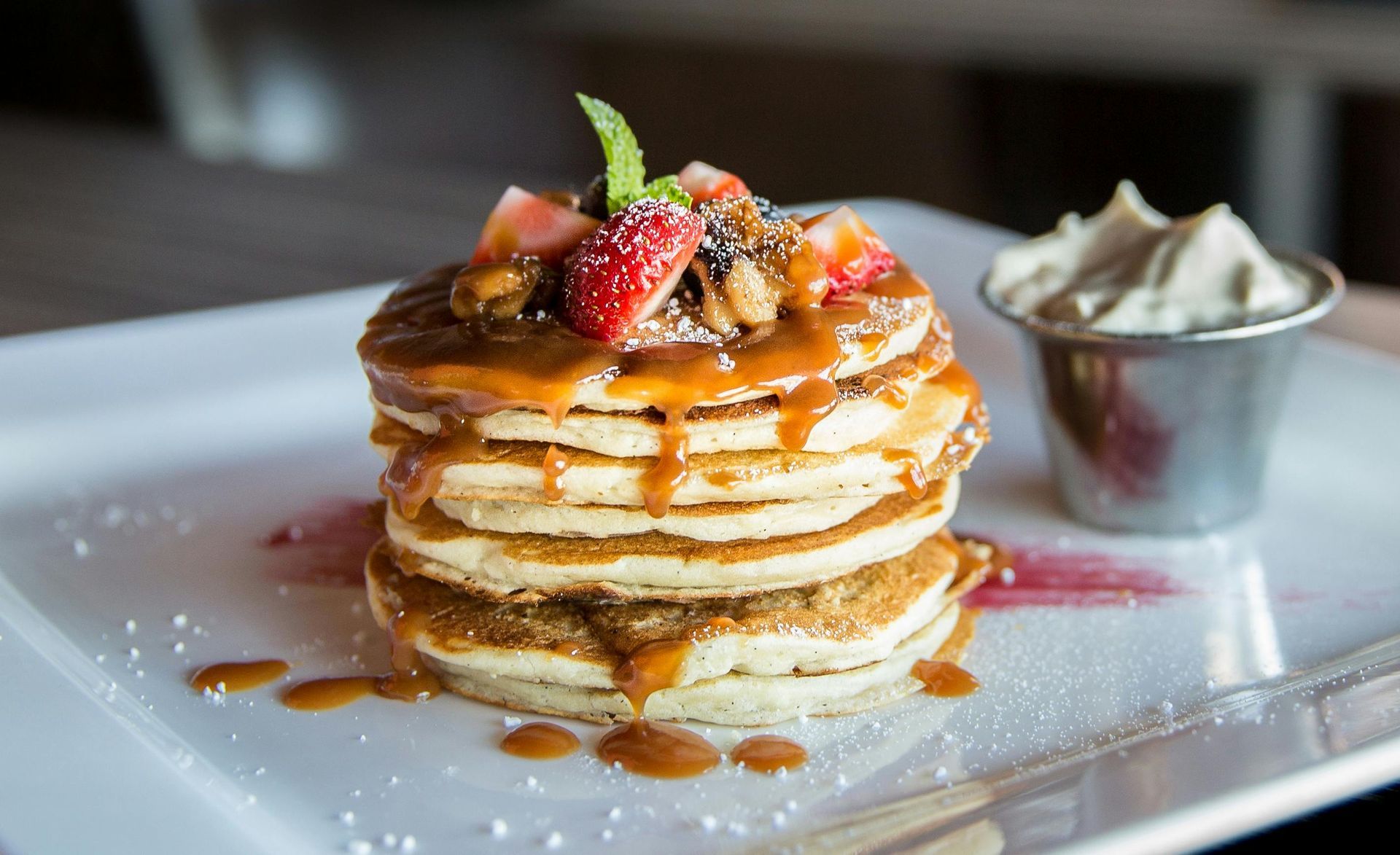 Stack of pancakes with caramel sauce, strawberries, nuts, and whipped cream on a white plate.