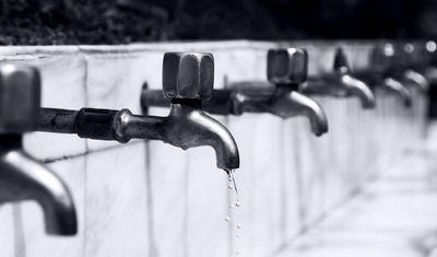 Row of metal water faucets, each with a small stream of water, against a white tiled wall.
