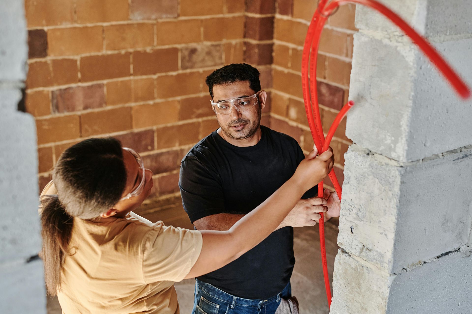 Two people installing red electrical wiring in a brick and concrete wall.