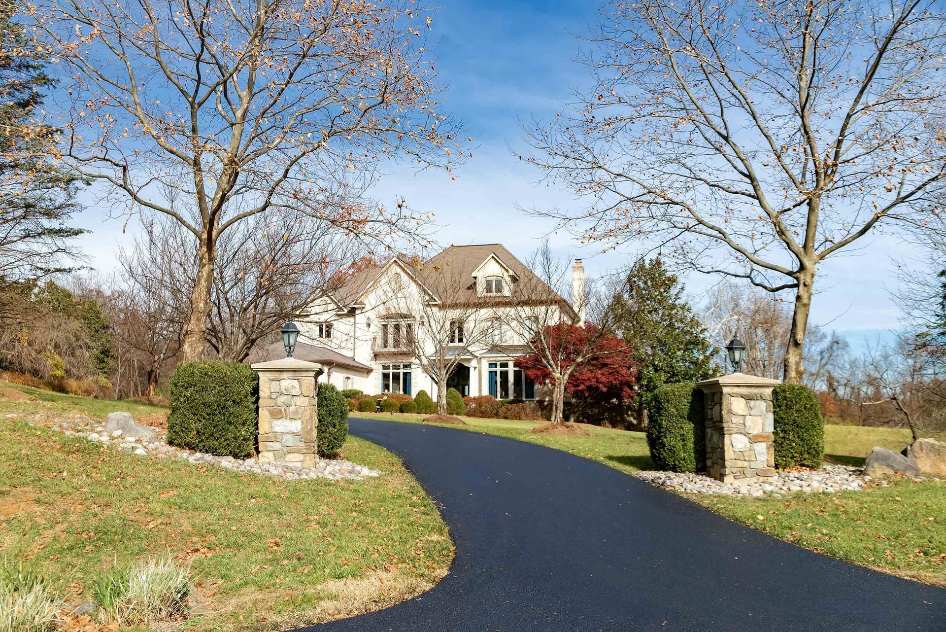 A large, two-story stone house at the end of a paved driveway, framed by trees and manicured bushes under a blue sky.