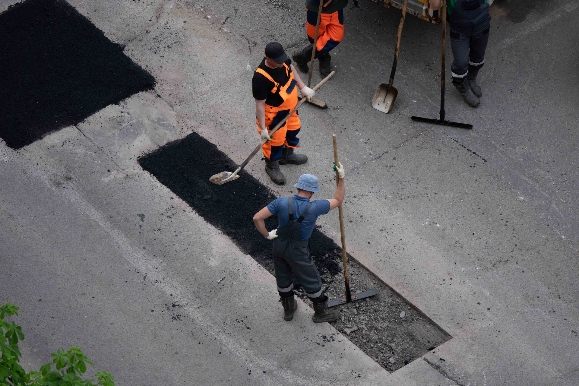 Construction workers in high-visibility gear paving a road with asphalt using shovels and a long-handled rake.