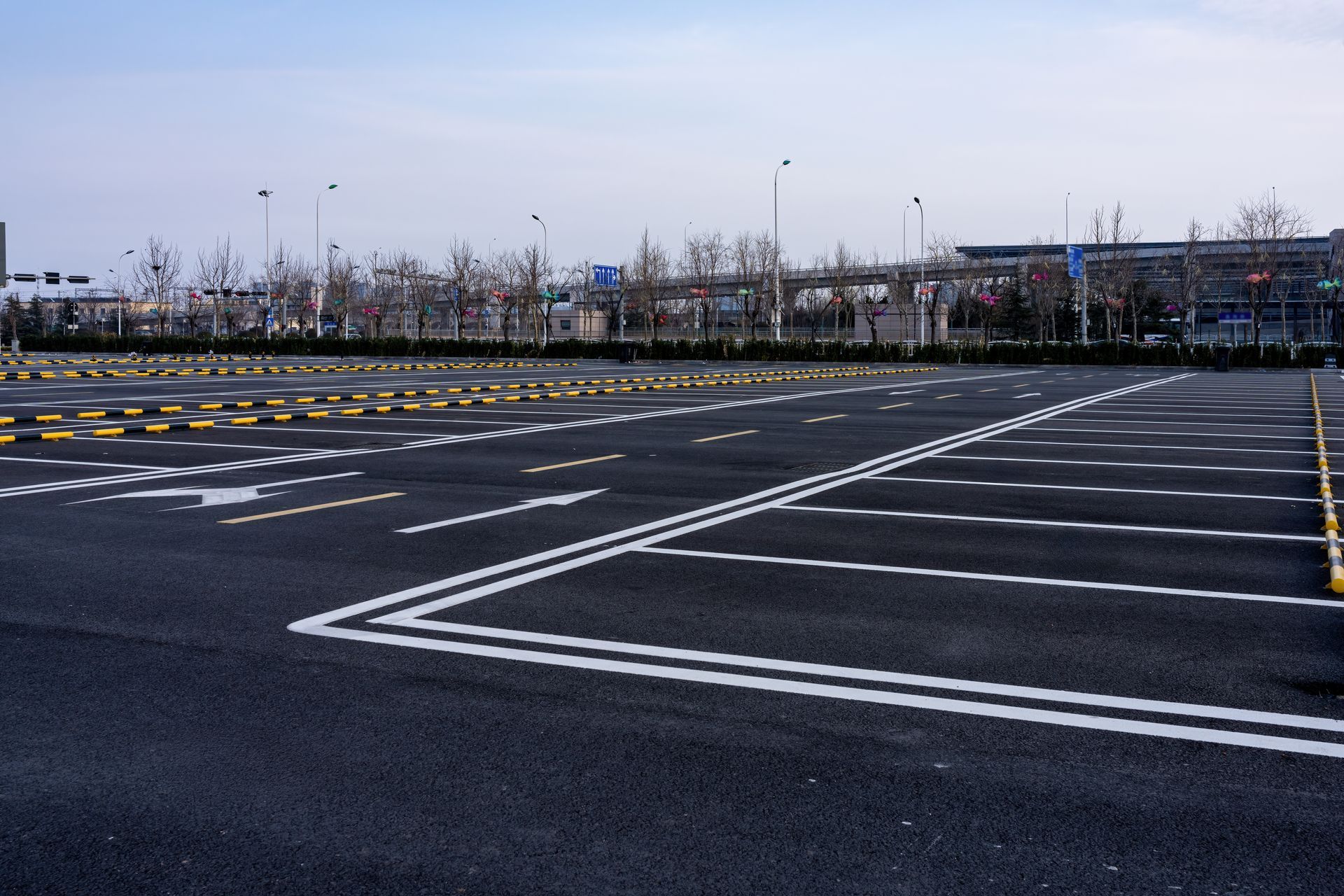 An expansive, empty asphalt parking lot featuring white lane markings and yellow parking barriers under a pale sky.