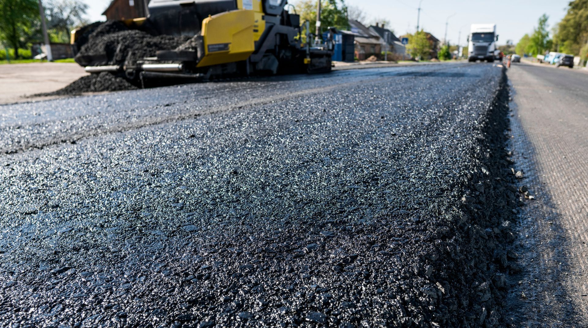 A yellow road paver machine lays a fresh layer of dark asphalt on a road during a sunny day.