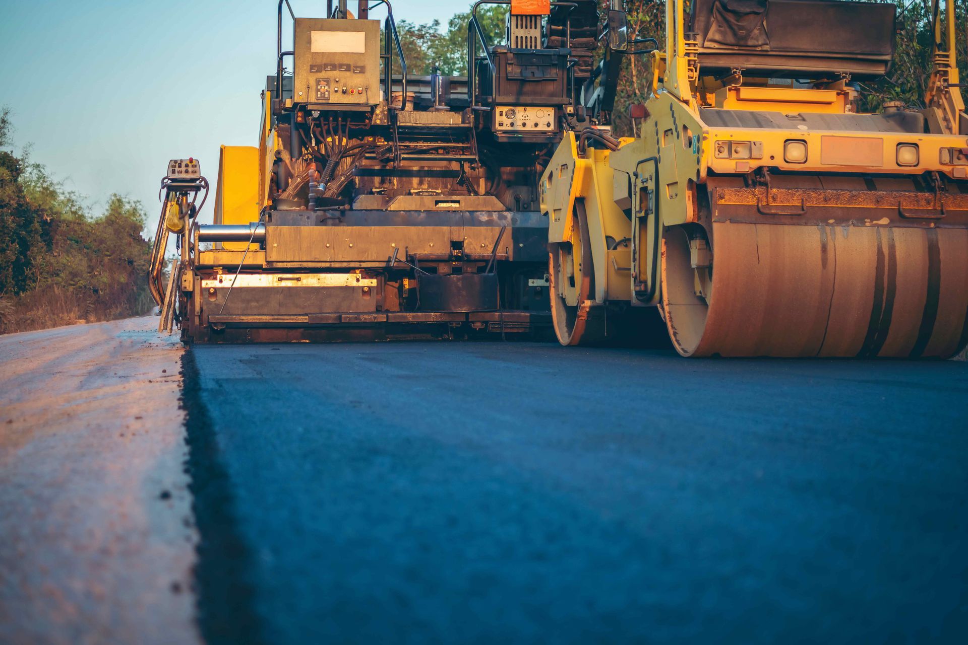 A bright yellow road paver and a smooth-drum roller operate on a paved road surface during a sunny day.