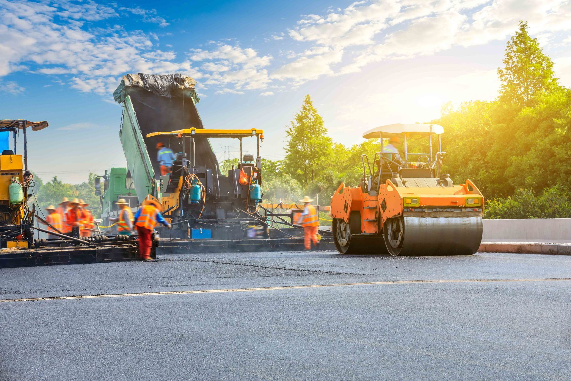 Workers pave a road with heavy machinery under a sunny sky, with a dump truck and a steamroller in operation.