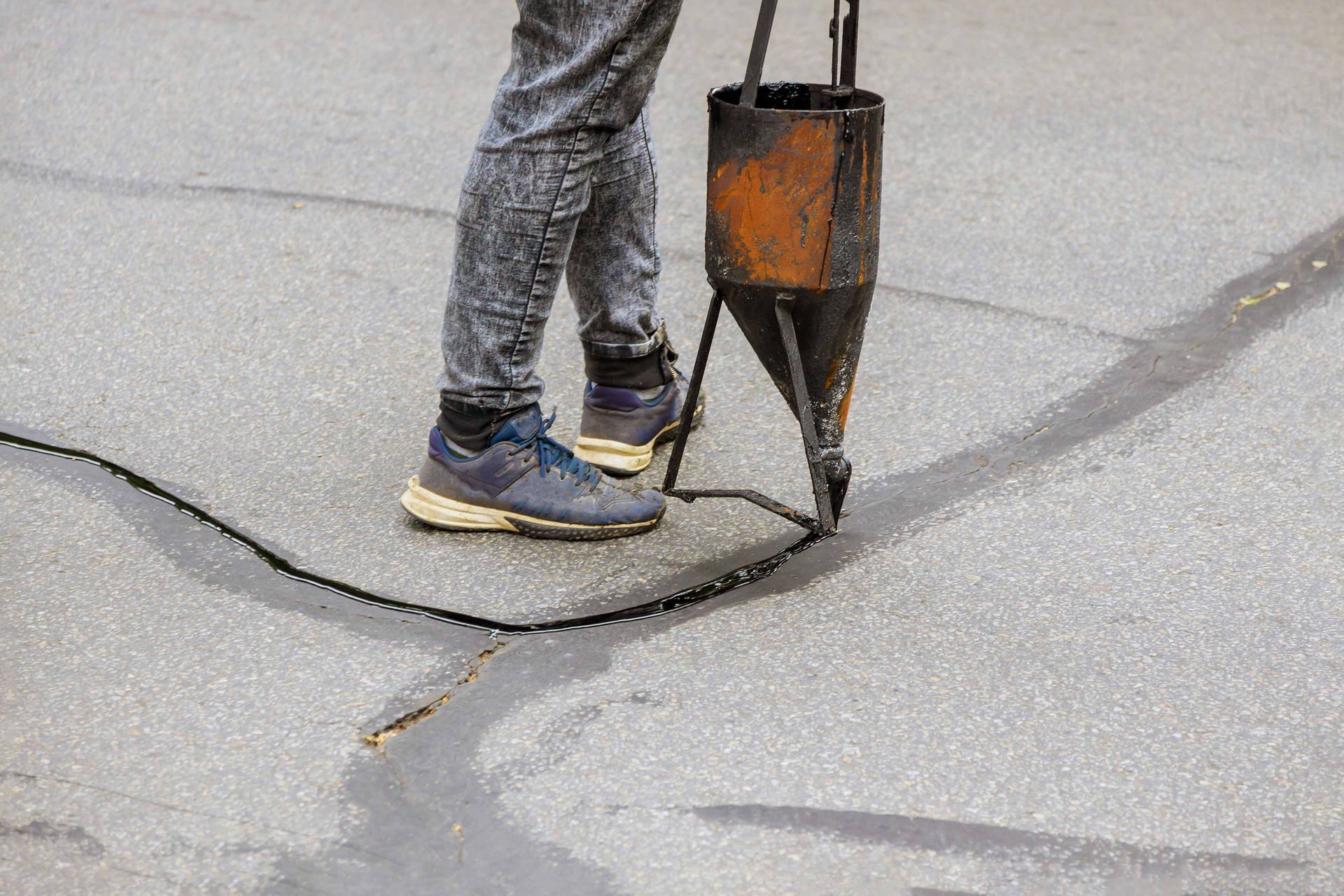 A worker uses a handheld pour pot to fill cracks in asphalt pavement with hot liquid sealant.